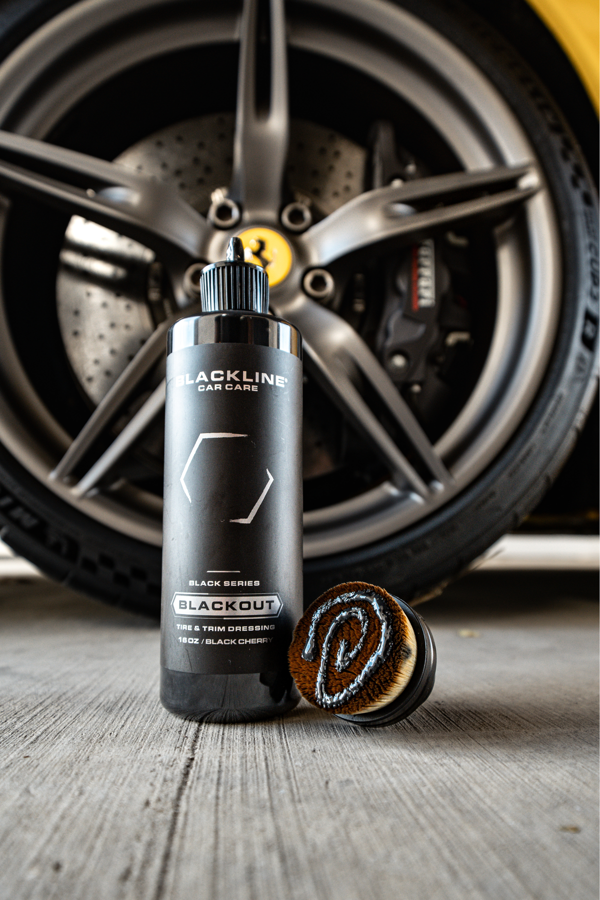 Yellow-themed Blackline Blackout trim and tire dressing bottle in front of a wheel with bronze rim and red brake caliper.
