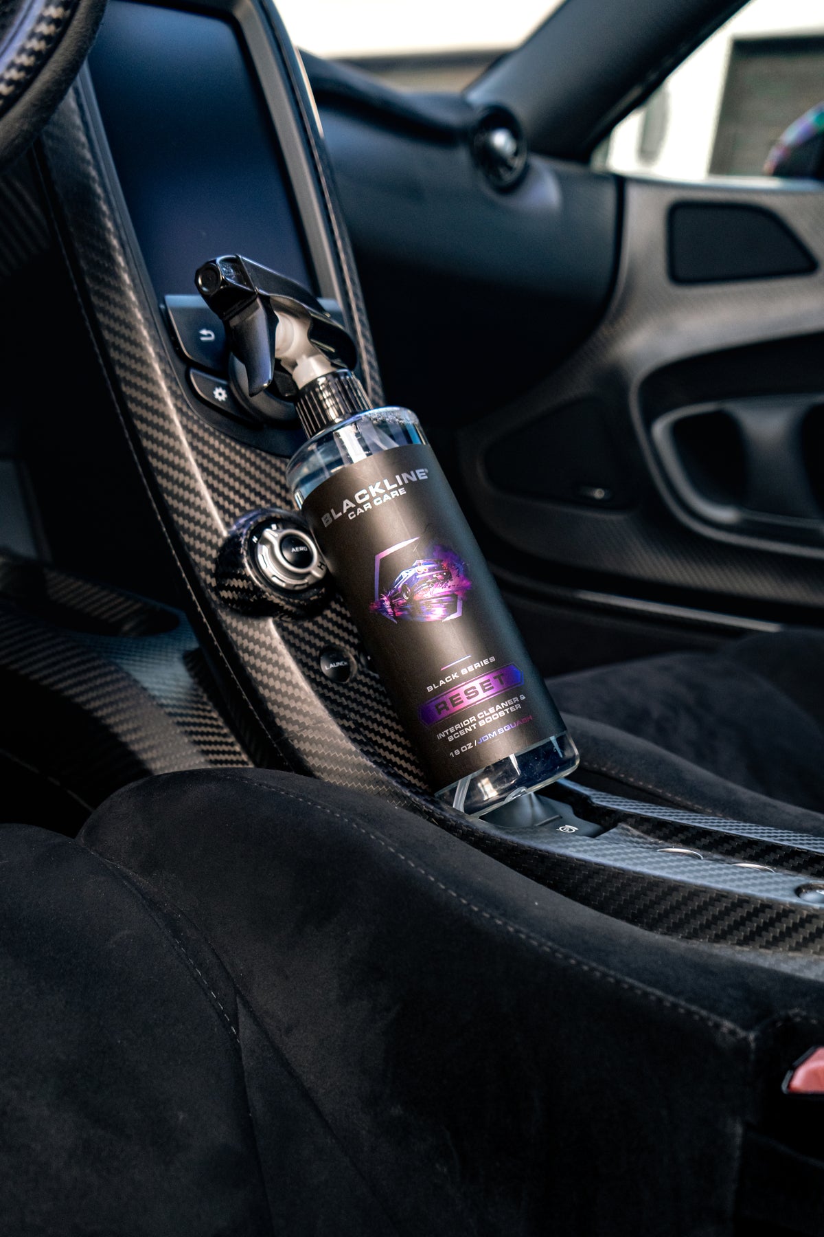 Blackline Reset interior cleaner JDM Squash scent bottle in a carbon-fiber vehicle interior, between two black sport bucket seats.